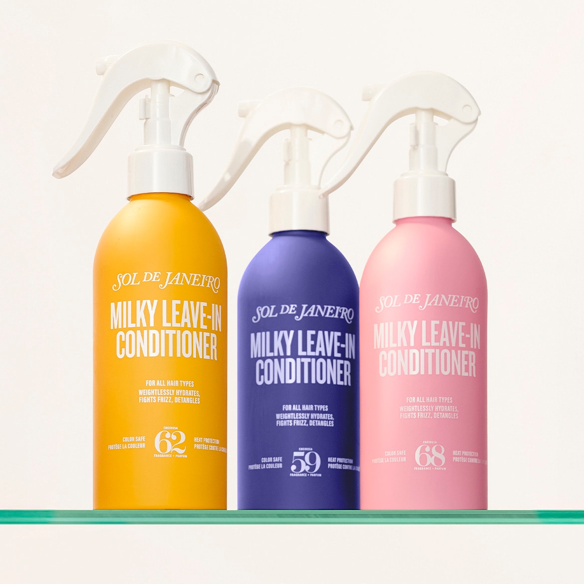 Drie flessen Sol de Janeiro Milky Leave-In Conditioner in geel, paars en roze, elk met een witte sproeikop, staan op een glazen plank en beloven hydratatie, frizz-bestrijding en ontwarring voor alle haartypes.
