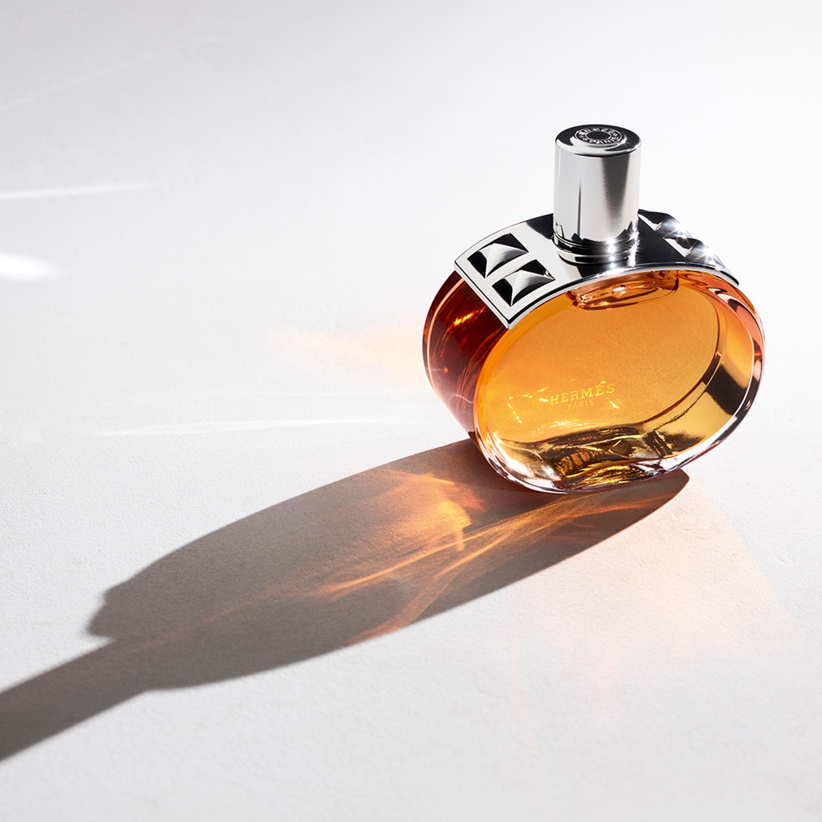 Een elegante, ronde parfumfles van Hermès met amberkleurige vloeistof en een zilverkleurige dop, die een lange schaduw werpt op een witte achtergrond.