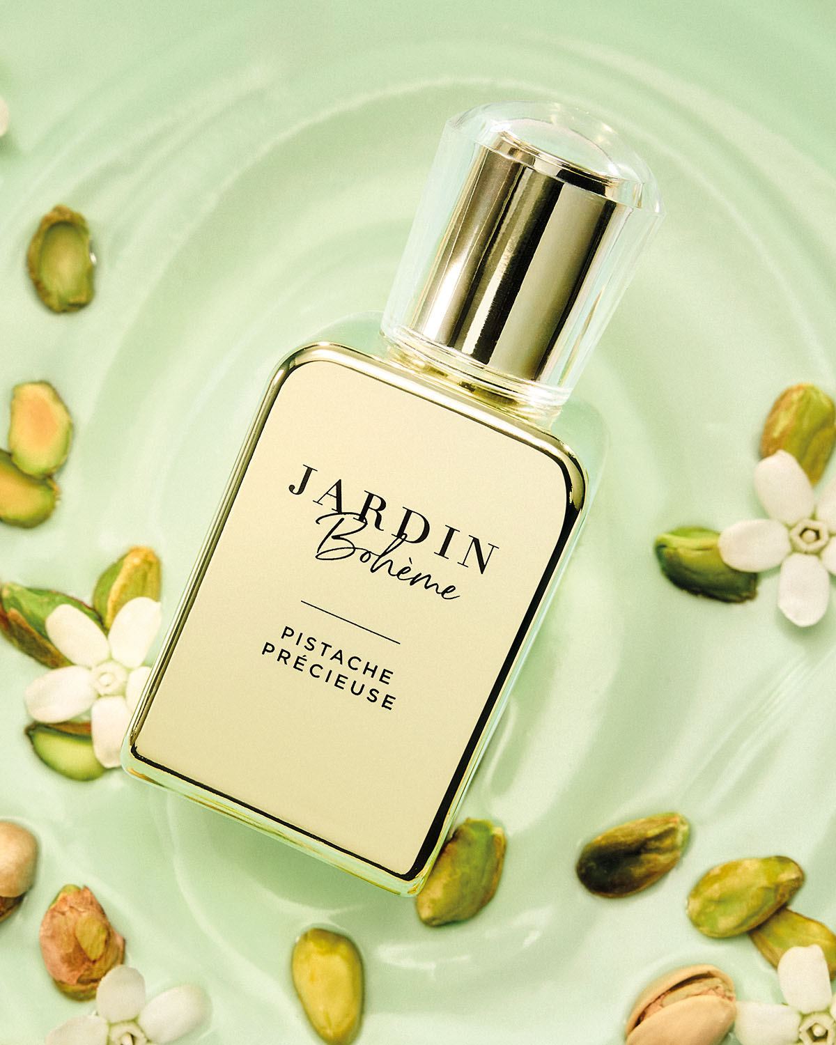 De Jardin Bohème Pistache Précieuse parfumfles, met een goudkleurige dop en een lichtgroen etiket, ligt op een lichtgroen oppervlak omringd door pistachenoten en witte bloemen.