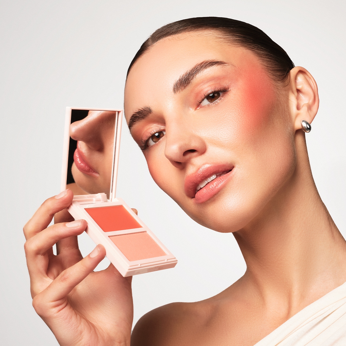 Een vrouw met een perzikkleurige blos op haar wangen en oogleden houdt een compacte blush met twee tinten vast, waarvan de spiegel haar lippen en neus reflecteert.
