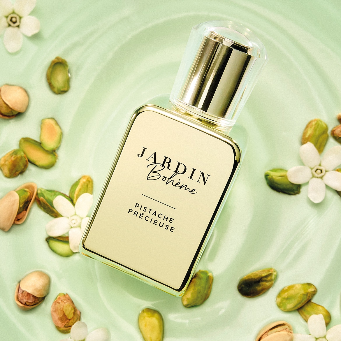 Jardin Bohème Pistache Précieuse parfumflesje met een gouden dop en een lichtgroen etiket, omringd door pistachenoten en witte bloemen op een lichtgroene ondergrond.