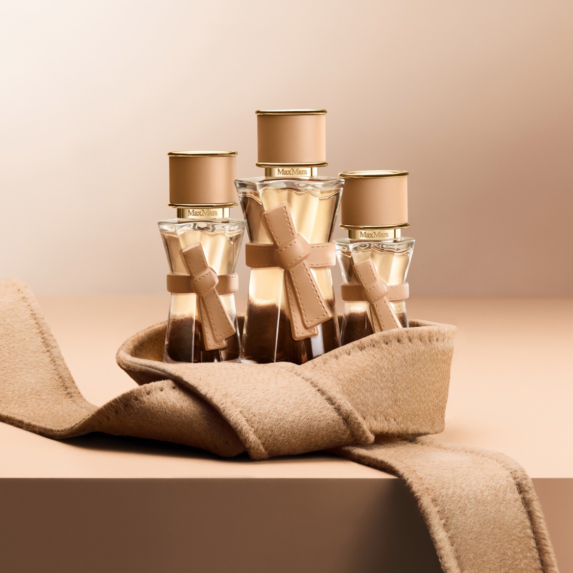 Drie Max Mara-parfums in elegante glazen flacons met goudkleurige doppen en beige strikjes, omringd door een zachte beige sjaal.