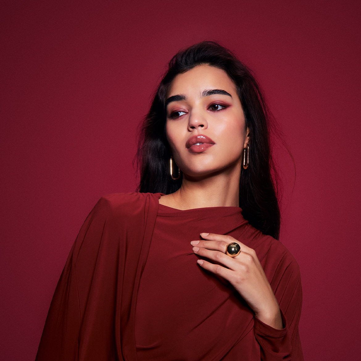 Een vrouw met lang, donker haar draagt een rode jurk, gouden oorbellen en een ring, met een rode oogschaduw en glanzende lippen.