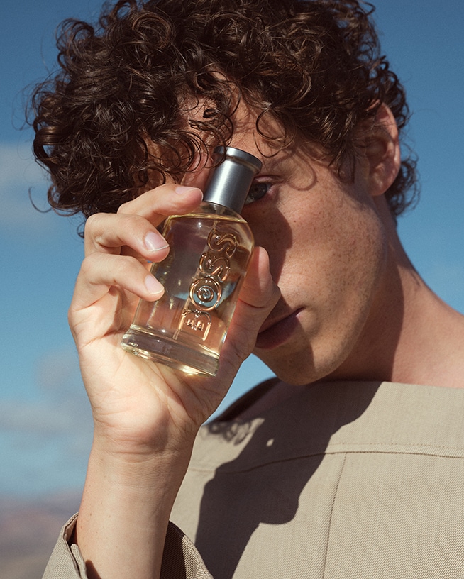 Jonge man met krullend haar houdt een heldere glazen fles Boss-parfum vast tegen een blauwe lucht.