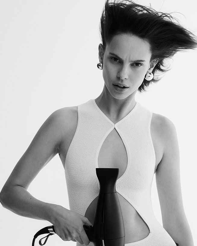 Een zwart-wit foto van een vrouw met kort, opgestoken haar, grote zilveren oorbellen en een witte top, die een zwarte föhn vasthoudt.