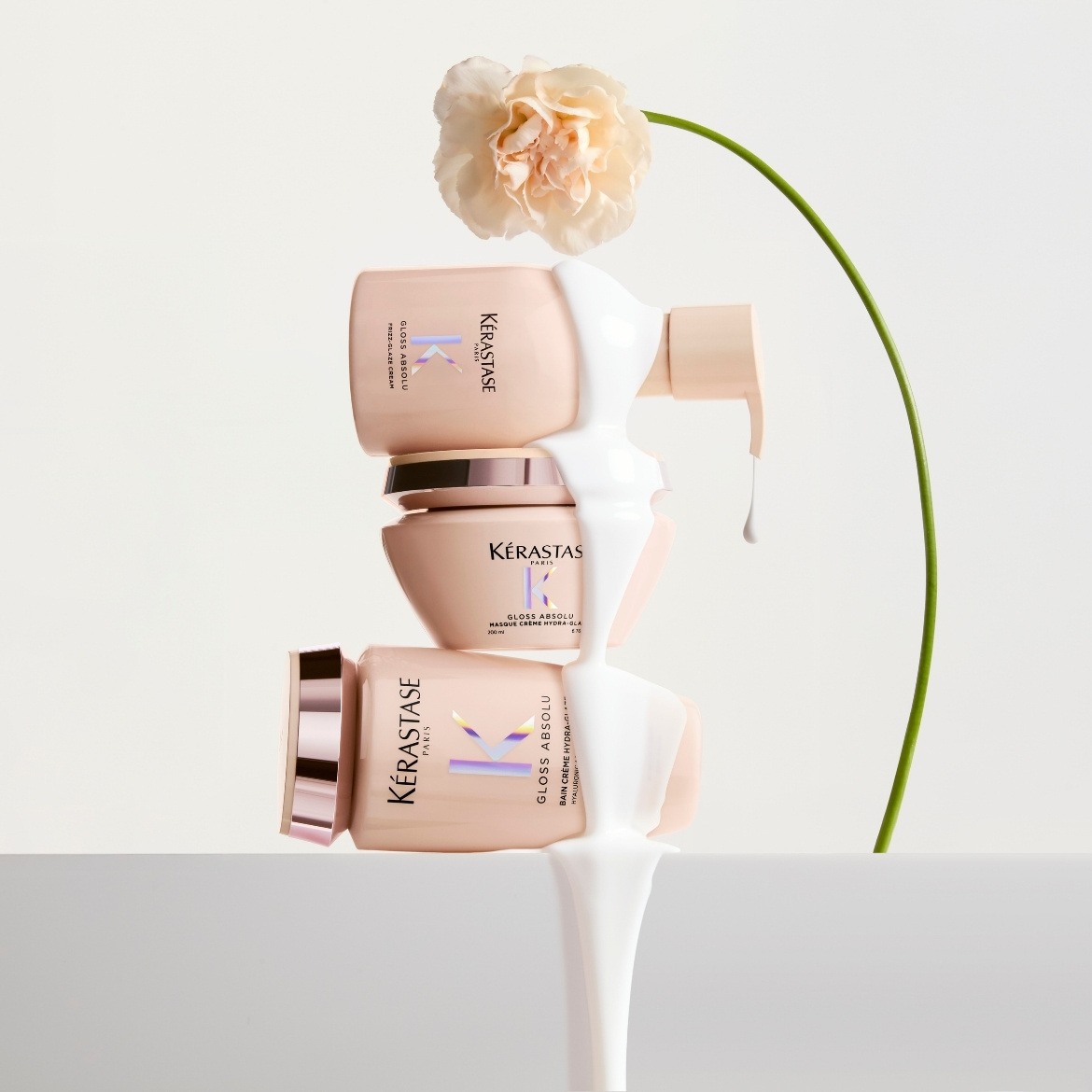 Stapel van Kérastase Gloss Absolu haarverzorgingsproducten in zachtroze verpakkingen, bedekt met witte crème, naast een delicate bloem.