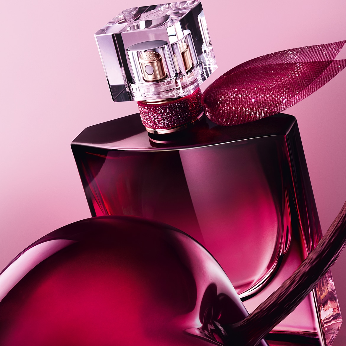 Een close-up van een Lancôme La Vie Est Belle-parfumflesje met een roze vloeistof, een glinsterende roze strik en een heldere dop, staand op een glanzend roze oppervlak.
