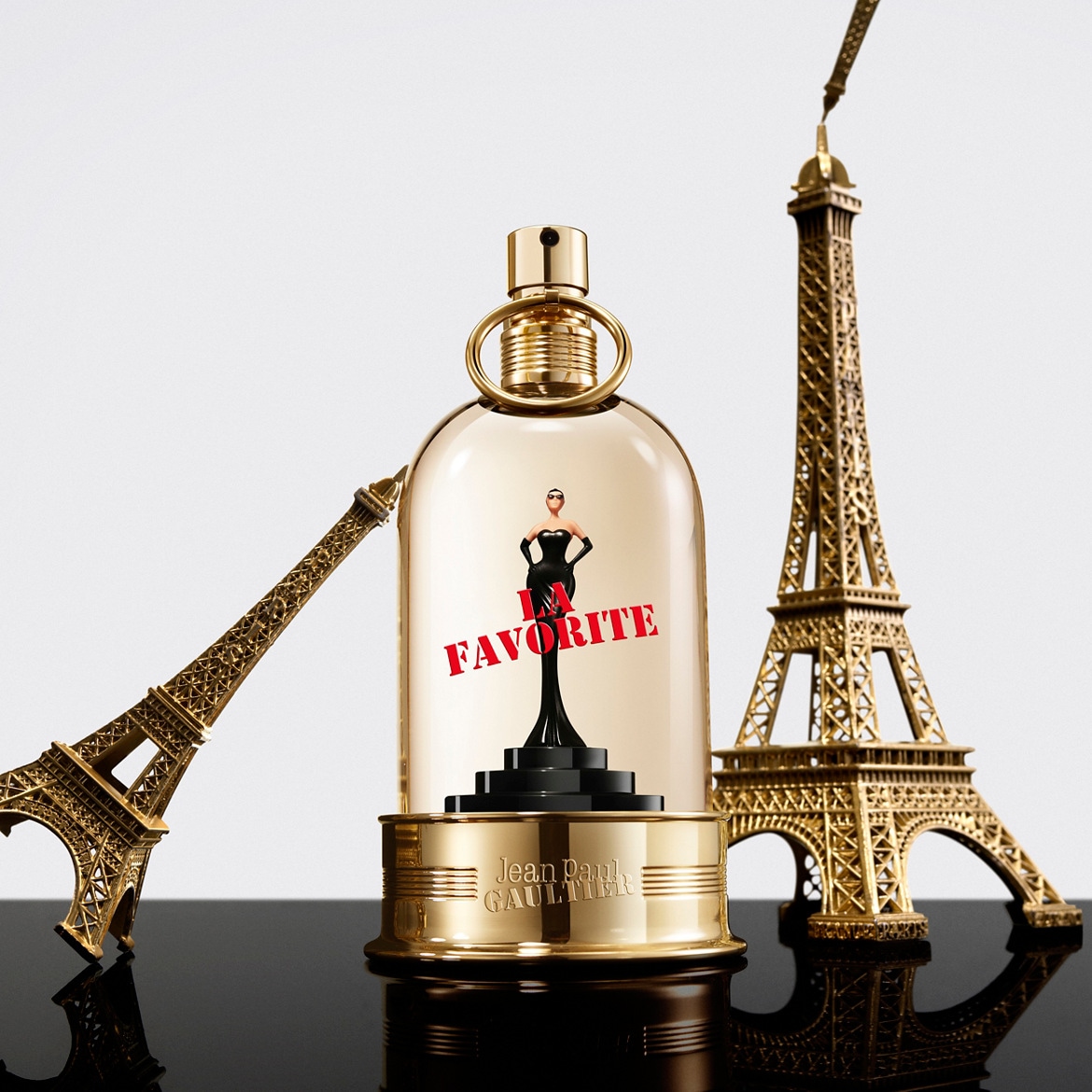 De Jean Paul Gaultier La Favorite parfumfles is een gouden trofee met een miniatuur van een vrouw in een zwarte jurk en de tekst "La Favorite" in rood, geflankeerd door twee gouden Eiffeltorens.