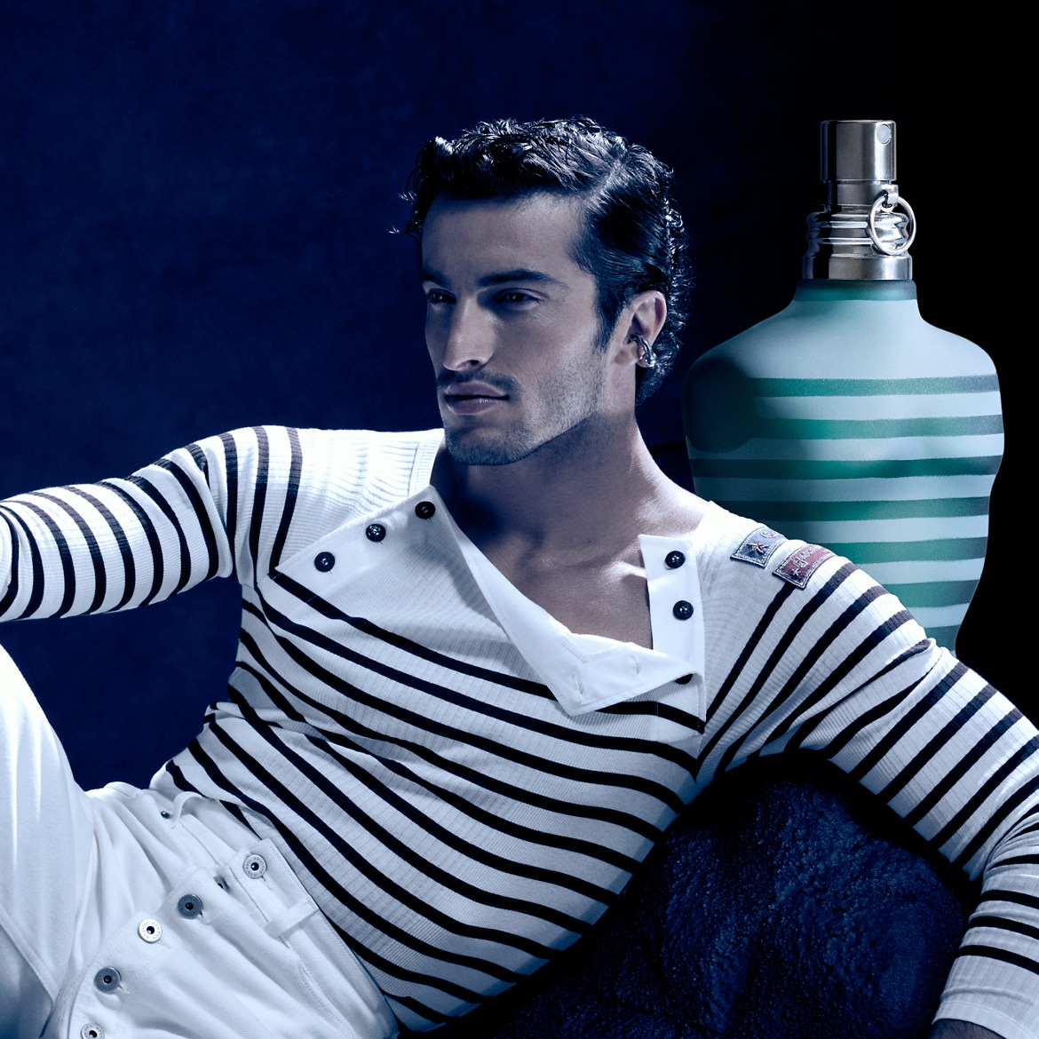 Een man poseert naast een fles Le Male Jean Paul Gaultier-parfum, gekleed in een gestreepte trui en witte broek, tegen een donkerblauwe achtergrond.