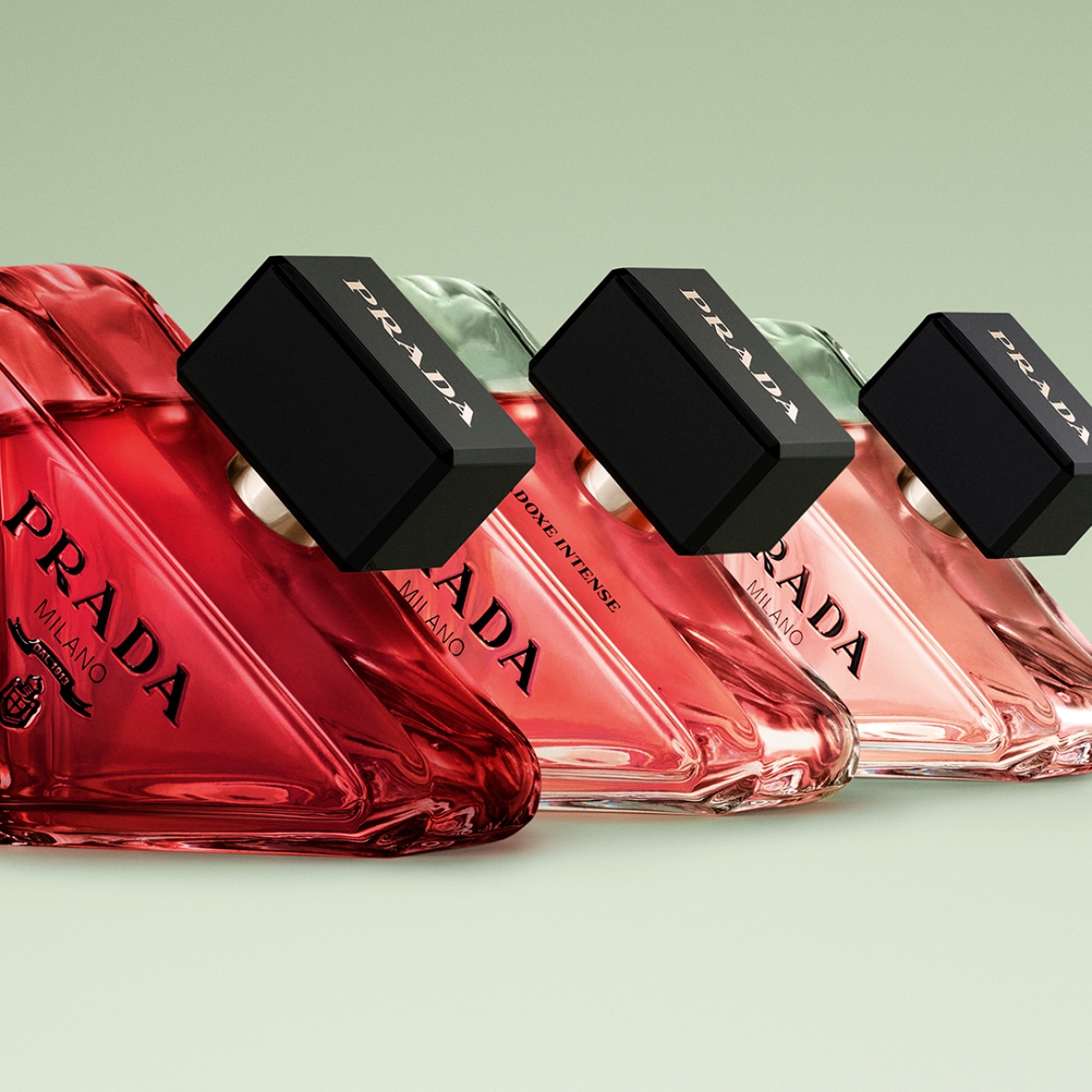 Vier Prada-parfums staan op een rij, elk met een driehoekige fles in verschillende tinten rood en roze, afgesloten met een zwarte dop met het Prada-logo.