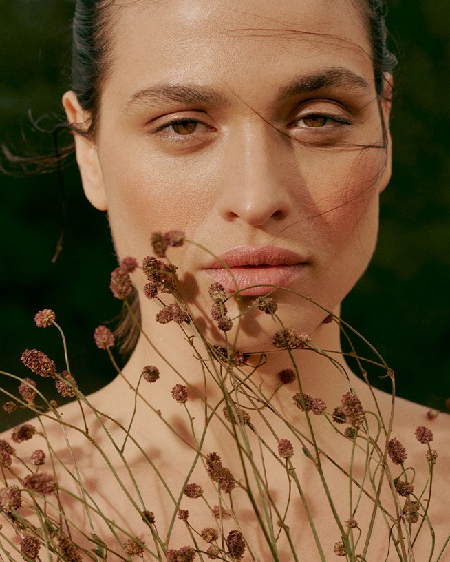 Een vrouw met een natuurlijke make-up look, omringd door kleine, bruine bloemen die haar gezicht omlijsten, waardoor een serene en aardse uitstraling ontstaat.